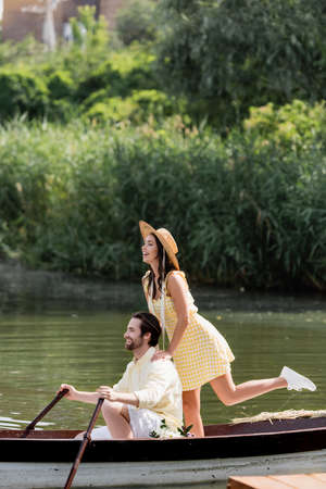 joyful young woman in straw hat standing behind man during romantic boat tripの写真素材