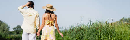 back view of young couple in summer clothes holding hands and standing near lake, bannerの写真素材
