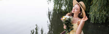 dreamy young woman adjusting straw hat and holding flowers near lake, banner