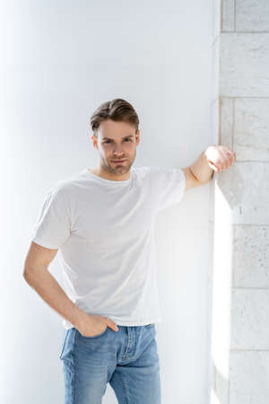 man in white t-shirt standing with hand in pocket of jeans near white wallの写真素材