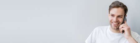 cheerful man in white t-shirt talking on mobile phone isolated on grey, banner