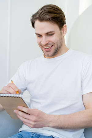 young and happy man in white t-shirt writing in diary at homeの写真素材