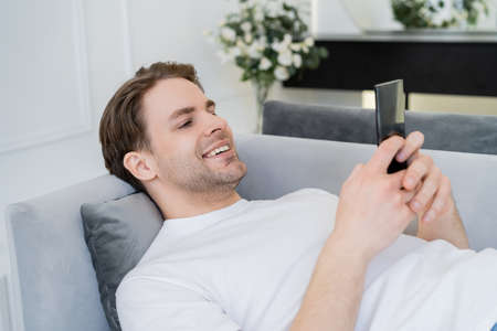 pleased young man resting on couch and chatting on smartphone