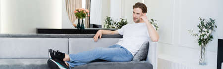 full length view of thoughtful man sitting on sofa in living room with floral decor, bannerの写真素材