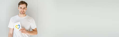 patriotic man holding card with blue and yellow heart and smiling at camera on grey background, bannerの写真素材