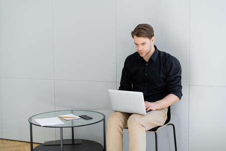 young man working on laptop while sitting near smartphone with blank screen on coffee table