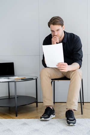 full length of thoughtful man looking at document while sitting near laptop with blank screenの写真素材