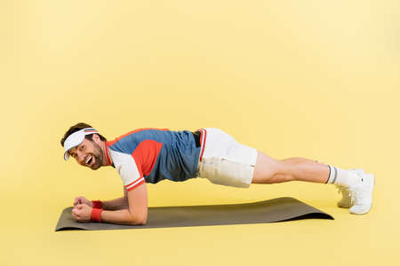 Positive sportsman standing in plank on fitness mat on yellow background