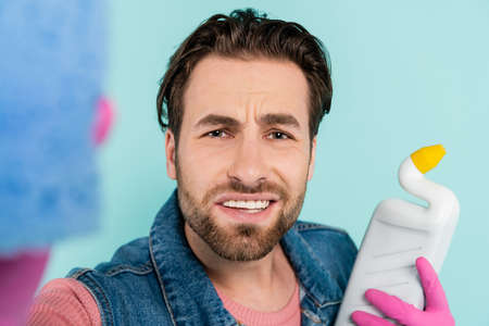 Young man in rubber gloves holding sponge and detergent isolated on blue