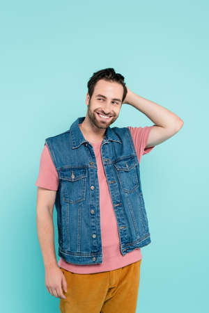 Smiling man in denim vest touching hair isolated on blueの写真素材