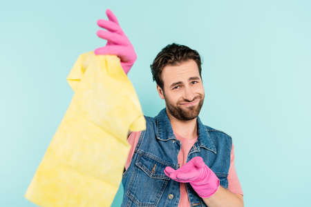 Smiling man in rubber gloves pointing at blurred rug isolated on blueの写真素材