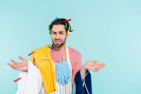 Displeased unmarried man in clothespins holding clothes isolated on blue