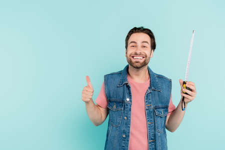 Positive man in denim vest holding measure tape and showing like isolated on blue
