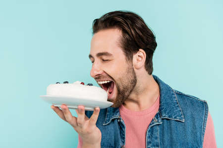Young man in denim vest opening mouth near cake isolated on blue