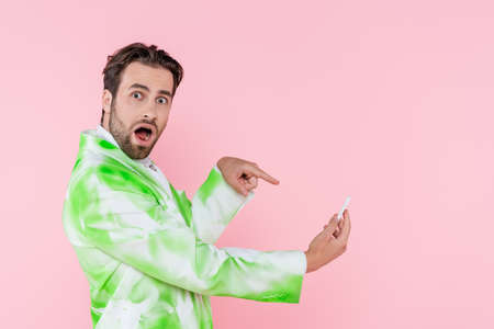 Young man in stylish jacket pointing at smartphone isolated on pinkの写真素材