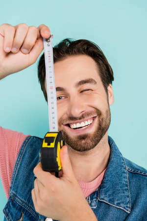 Smiling man in denim vest holding blurred tape measure isolated on blueの写真素材