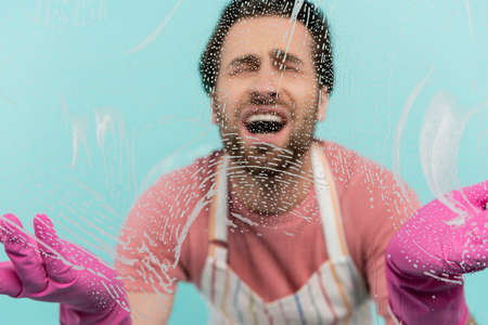 Blurred upset man in rubber gloves standing near glass with detergent isolated on blue