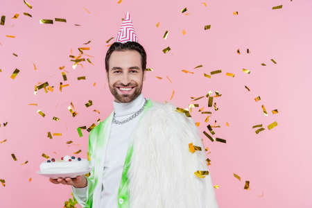 Positive man in furry jacket and party cap holding birthday cake under confetti on pink background
