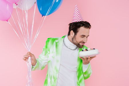 Young man in party cap smelling birthday cake and holding balloons isolated on pinkの写真素材
