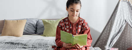 preteen girl reading book while sitting on bed near pillows and wigwam, banner