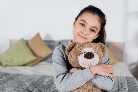 cheerful preteen girl embracing teddy bear and looking at camera in bedroomの写真素材