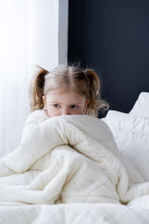 scared girl sitting on bed and obscuring face with white blanketの写真素材