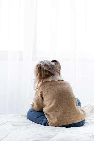 back view of little girl with ponytails sitting alone on bed at homeの写真素材