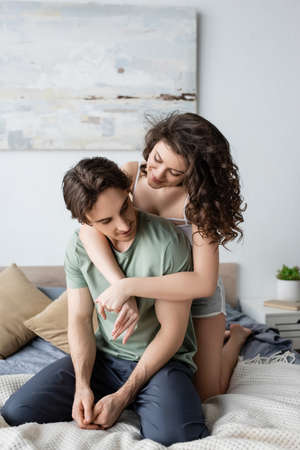 pleased and curly woman hugging boyfriend in bedroomの写真素材