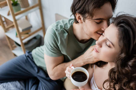high angle view of happy man kissing curly girlfriend with cup of coffeeの写真素材