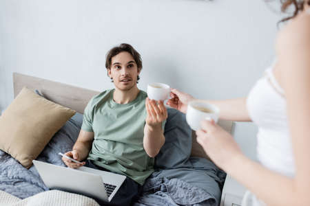 blurred woman holding cups of coffee near boyfriend using laptop in bedの写真素材