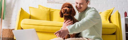 Positive teenager with down syndrome pointing with finger at laptop and hugging poodle at home, bannerの写真素材