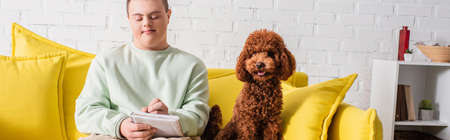 Teen boy with down syndrome writing on notebook near poodle on couch, banner
