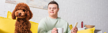 Teen boy with down syndrome holding cup and smartphone near poodle at home, banner