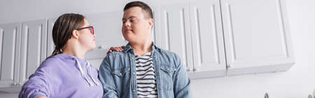 Low angle view of teenager with down syndrome looking at boyfriend in kitchen, bannerの写真素材