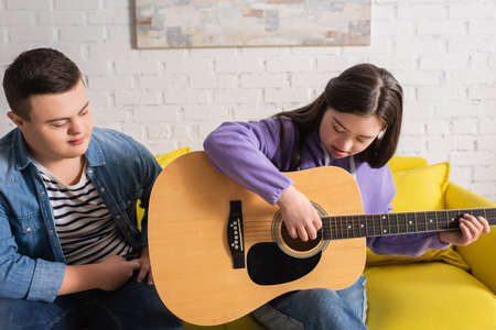 Girl with down syndrome playing acoustic guitar near friend on couch at homeの写真素材