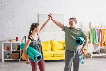 Positive teenagers with down syndrome holding fitness mats and giving high five at home