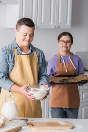 Smiling teenager with down syndrome holding flour near blurred girlfriend with baking sheet in kitchenの写真素材