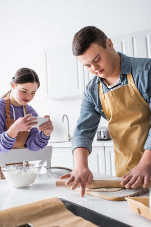 Teen boy with down syndrome rolling dough near friend with smartphone in kitchenの写真素材