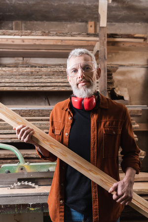 bearded woodworker with board looking at camera in workshopの写真素材