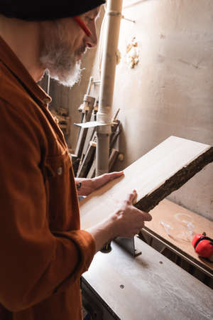 bearded woodworker in beanie holding board in workshopの写真素材