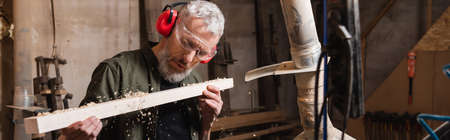 woodworker in goggles blowing sawdust from plank, banner