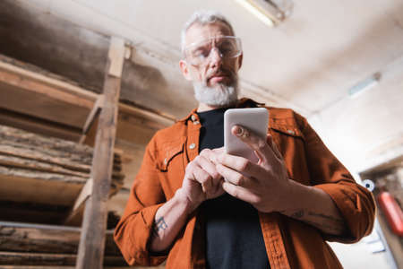 low angle view of bearded furniture designer using smartphone in woodwork studioの写真素材