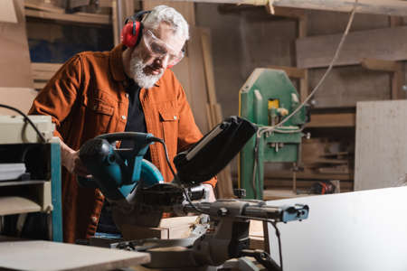 bearded joiner in goggles working on miter saw in workshopの写真素材
