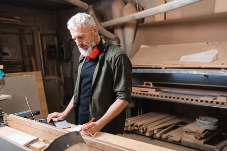 middle aged furniture designer writing near laptop on workbenchの写真素材