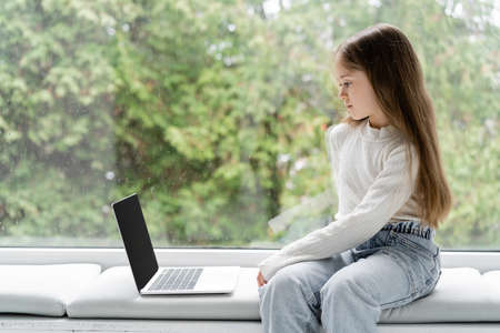 girl sitting on windowsill at home near laptop with blank screenの写真素材
