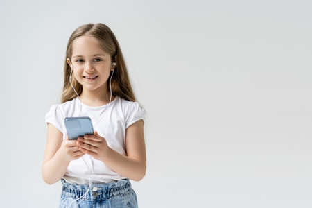 happy child with earphones and cellphone looking at camera isolated on grey