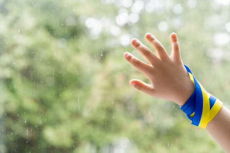 cropped view of child with blue and yellow ribbon touching windows glass with raindropsの写真素材