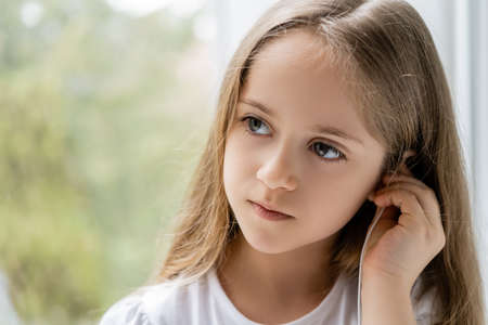 girl with long hair adjusting earphone near blurred window