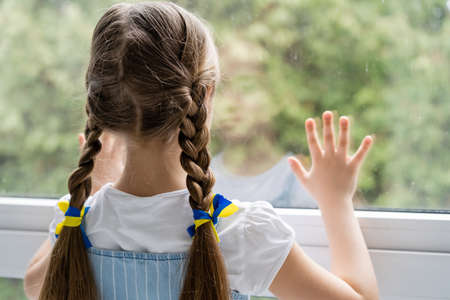 back view of girl with blue and yellow ribbons on braids standing near window at homeの写真素材