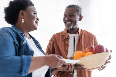 happy senior african american man holding bowl with fresh fruits near smiling wifeの写真素材